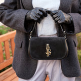 Person wearing a gray coat and black gloves holding a black Carlo Salvatelli handbag in suede with a gold emblem.