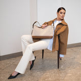 Woman in a brown coat and white pants holding a beige handbag in an indoor setting.