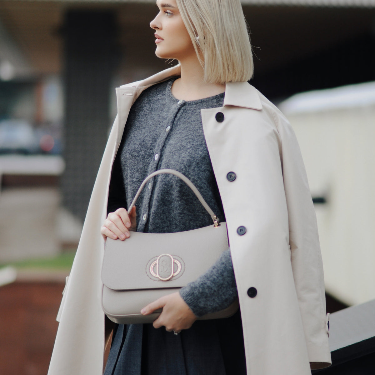 Woman holding a beige handbag wearing a long coat and dress.