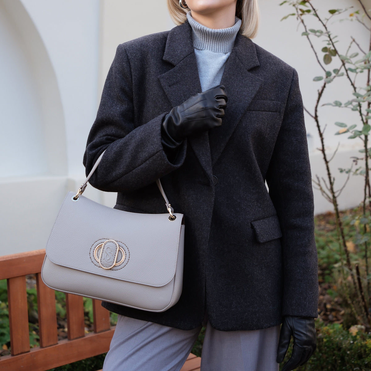 Woman in a dark coat holding a light gray handbag outdoors.