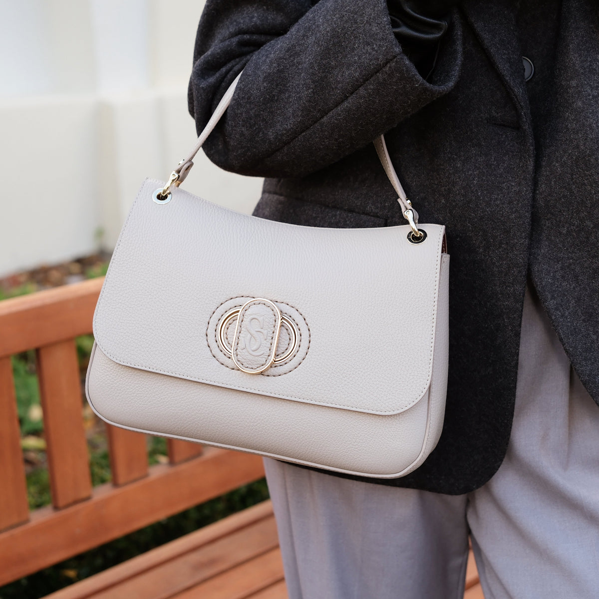 Person holding handbag with a Carlo Salvatelli logo, wearing a dark coat and light pants, outdoors.