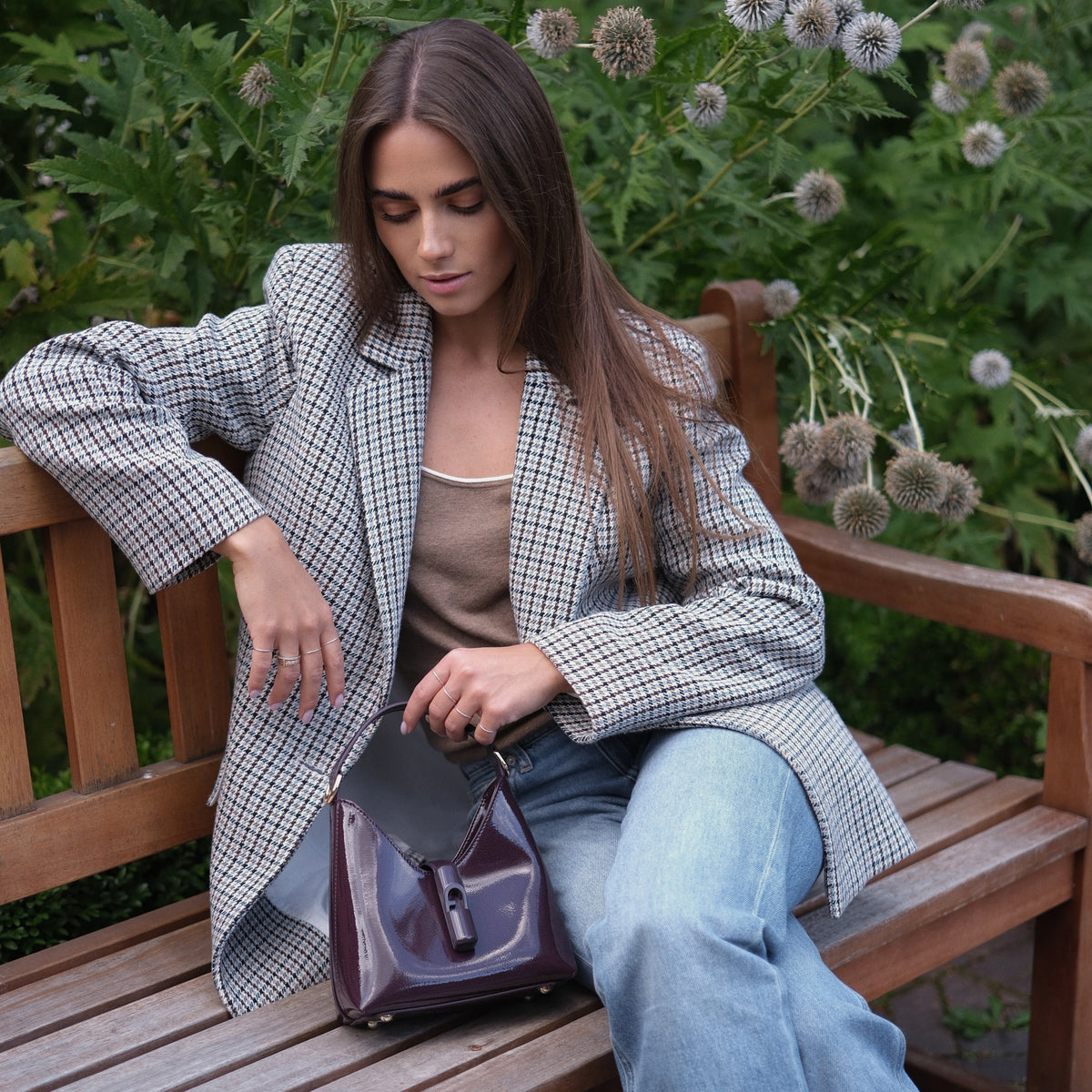 A woman sits on a wooden park bench wearing a black and white houndstooth blazer, a beige tank top, and light blue jeans, holding a dark purple handbag. The background features green foliage with dried thistle flowers.