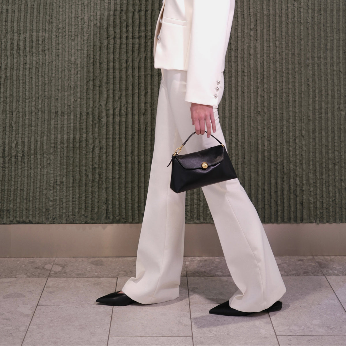 Person wearing a white outfit with a black handbag against a green textured wall.