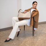 Woman in a brown coat and white pants holding a beige handbag in an indoor setting.