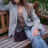 A woman sits on a wooden park bench wearing a gray and black checkered blazer, a brown top, and light blue jeans, with a dark purple handbag resting on the bench. Green foliage with dried flowers is in the background.