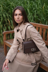 A woman wearing a beige trench coat sits on a wooden bench outdoors, carrying a dark brown structured shoulder bag with a chain strap and a silver-tone floral clasp.