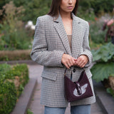 A woman standing in a garden wearing a black and white checkered blazer, light blue jeans, and holding a dark purple handbag.