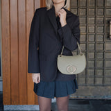 Woman in a black blazer and blue pleated skirt holding a green handbag against a wooden and tiled wall.