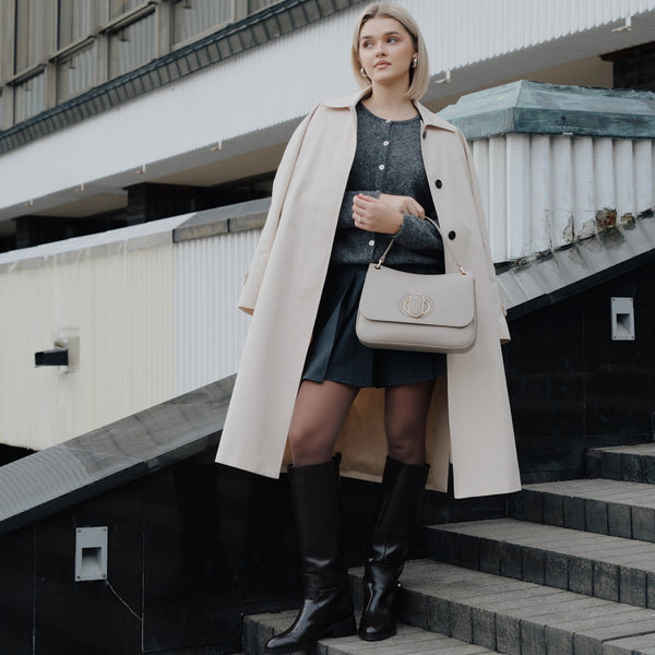 Woman in a beige coat and beige handbag standing on steps with a building in the background