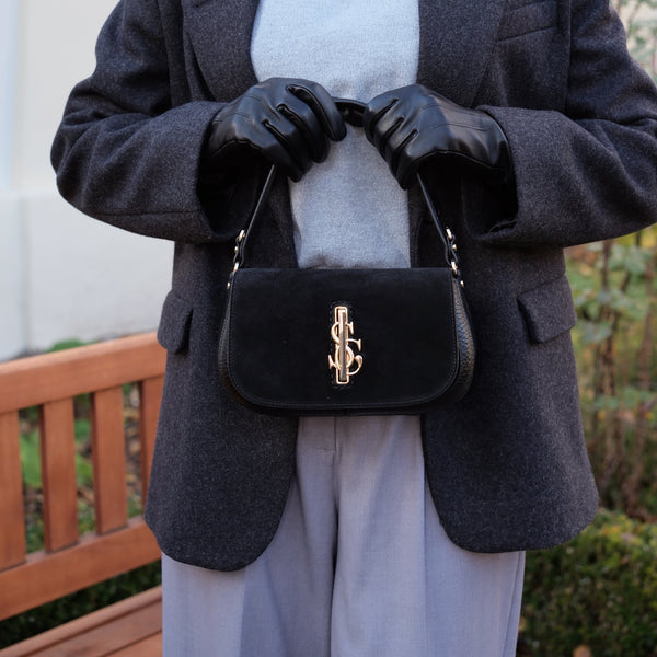 Person wearing a dark gray blazer, light gray turtleneck, and black gloves, holding a black Carlo Salvatelli handbag with a gold clasp.