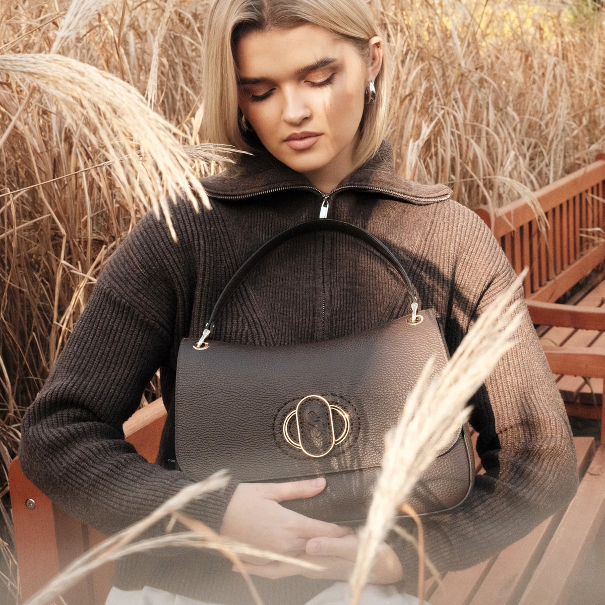Woman holding a black Carlo Salvatelli handbag in an outdoor setting with dry grass and a bench.