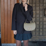 Woman in a black blazer and blue pleated skirt holding a green handbag against a wooden and tiled wall.