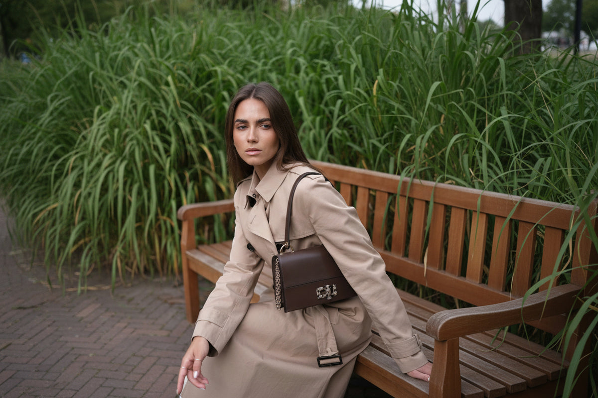 A woman wearing a beige trench coat sits on a wooden bench outdoors, carrying a dark brown structured shoulder bag with a chain strap and a silver-tone floral clasp.